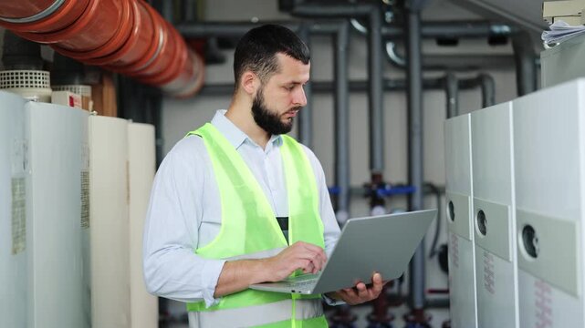 Focused engineer, bearded and wearing light blue striped shirt and high-visibility vest, actively uses laptop for data input and analysis amidst industrial pipes machinery within boiler room setting.