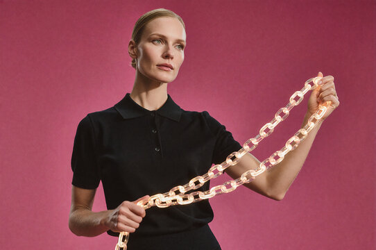 Woman holds an illuminated translucent chain against a magenta background. Soft studio light highlights her facial structure and the glowing amber links of the accessory.