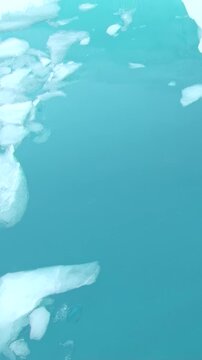 Vertical Video of Floating Ice Fragments Seen Through Blue Water. Broken white ice spreads across blue-green water, showing varied frozen shapes, texture and gentle ripples.
