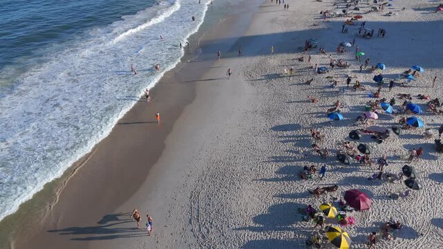 Aerial flyover of Copacabana Beach with beachgoers, colorful umbrellas and waves rolling onto the sand on a sunny day, Rio de Janeiro, Brazil.