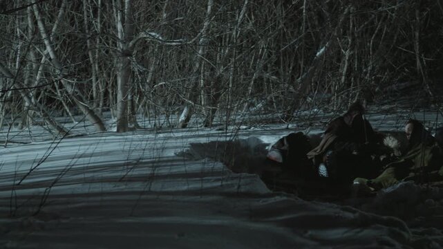 Two people rest on snowmobile in heavy winter clothing at night surrounded by dense forest and bare branches. scene is dimly lit with cool tones emphasizing cold environment
