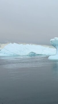 Vertical Video of White Iceberg Shelf Across Dark Water. White and blue ice, rippling water and soft diffuse light reveal natural frozen texture and sculpted contours across the marine scene.