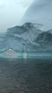 Vertical Video of Textured Iceberg Wall With Red-Hooded Passenger Below. A red hooded passenger appears at the lower frame edge while the massive carved ice face dominates the dark rippling water.