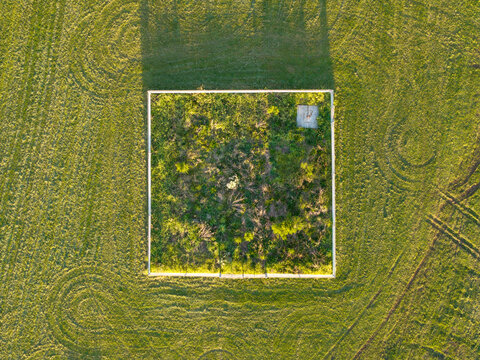 Aerial view of a square fenced enclosure containing wild vegetation set within a grassy field with visible tire tracks in Melbourne, VIC, Australia.