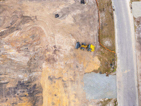 Aerial view of a yellow front-end loader on a dirt construction site with tire tracks and a paved road in Leland, North Carolina, United States.