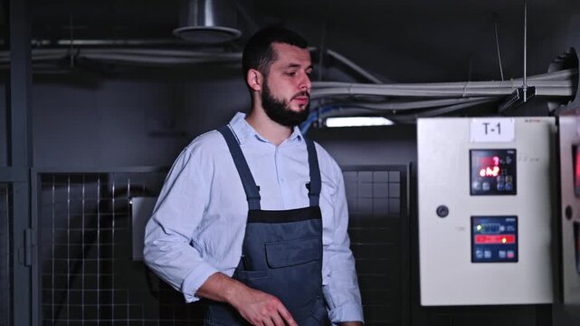 Puzzled engineer in work overalls ponders problem at control panel in dark technical room.