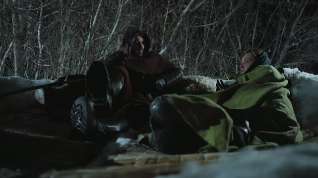 Two people rest on sled covered by snow at night within dense leafless trees. One person lies wrapped in green blanket other sits with hooded cloak and black gloves. Dim lighting creates