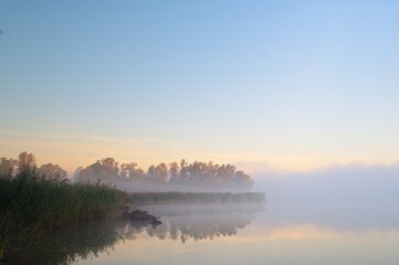 Delikatne, eteryczne ujęcie jeziora w porannej mgle z ciepłym pastelowym niebem, szuwarami i spokojnym odbiciem światła na powierzchni wody. © TomashRyś