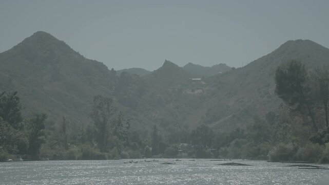 Hazy mountain peaks above green trees and a dark riverbed in soft light