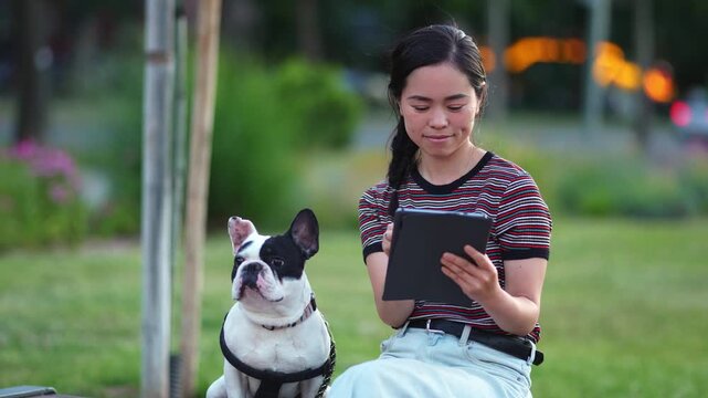 Cheerful japanese woman and her French Bulldog resting on a bench and using a digital tablet