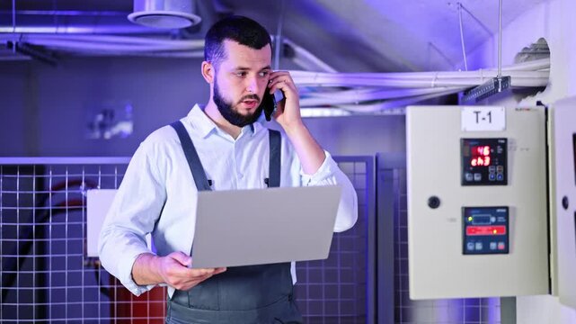 Diligent engineer, man with beard wearing light shirt and dark overalls, talking on phone while holding a laptop. Monitoring systems in modern server room, checking data and solving technical issues.