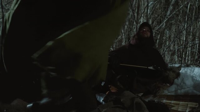 Hooded hunter sits quietly by snow covered ground holding arrow laden bow under dim light in dense winter woods. Another figure moves through scene partially obscured and interacting