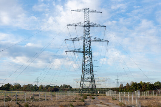 High voltage transmission tower rises at power line construction site with suspended conductors and fencing. Steel lattice structure represents grid expansion and energy engineering under cloudy sky