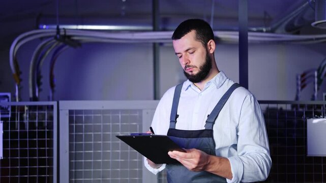 Diligent technician with beard and overalls writes on a clipboard, inspecting systems in a modern industrial data center. Focus on maintenance and technical documentation.