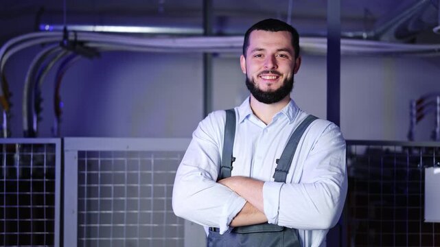 Smiling engineer with a beard, wearing a light blue shirt and work overalls, stands confidently with arms crossed in an industrial facility.