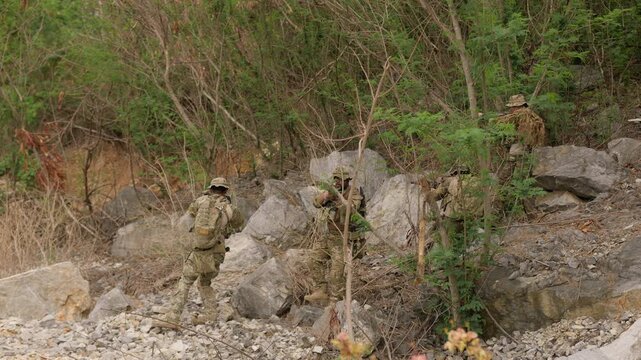 A team of soldiers in full combat gear conducts a tactical patrol, moving cautiously with rifles through a rugged, rocky landscape during a military operation.