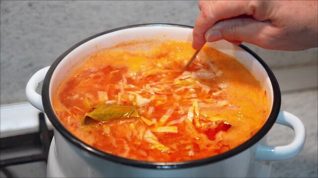 Cook stirring a hot red borscht or cabbage soup with a spoon in an enamel pot on a stove. Preparing traditional ethnic beetroot broth meal vegetable ingredients homemade culinary recipe process.
