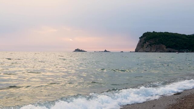 Sunset coastal wide view with gentle waves breaking on pebble beach, distant rocky headland, small islands, and calm horizon under pastel sky with soft evening glow
