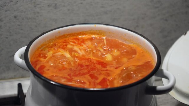 Hot red borscht soup bubbling and boiling in a white enameled pot on a stove indoors. Beautiful cinematic culinary food scene of a traditional homemade cabbage beetroot soup cooking process. 