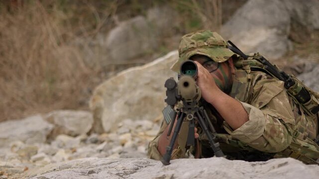 A special forces soldier in camouflage lies in a prone position, aiming his sniper rifle with a scope during a tactical surveillance mission in a rocky, arid terrain.