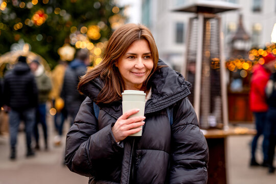 Smiling woman holding hot coffee in festive Christmas market lights during winter evening