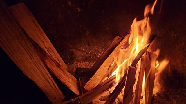 Flames leap over stacked wood logs in dark setting. Warm light dances on charred timber surface. Fires crackle, casting golden glow on surrounding darkness