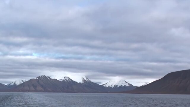 Arctic Ocean Ship Navigating Cold Arctic Ocean Under Northern Sky. Snow-covered mountains remain visible beyond the cold open water and the distant polar horizon.