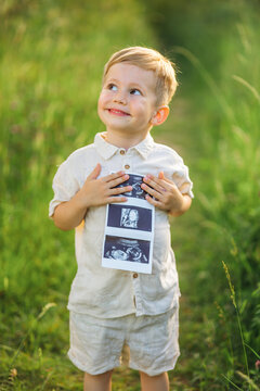 Little boy proudly holds ultrasound photos in a sunny meadow, announcing a new sibling