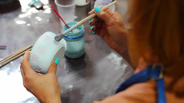 Woman applying blue glaze to handmade ceramic mug with brush. Pottery glazing process represents ceramic craft and handmade production