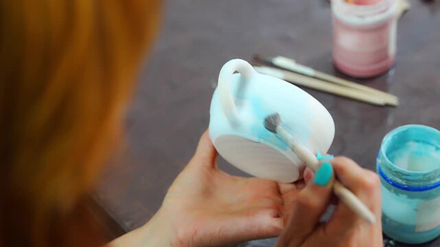 Woman applying blue glaze to handmade ceramic mug with brush. Pottery glazing process represents ceramic craft and handmade production
