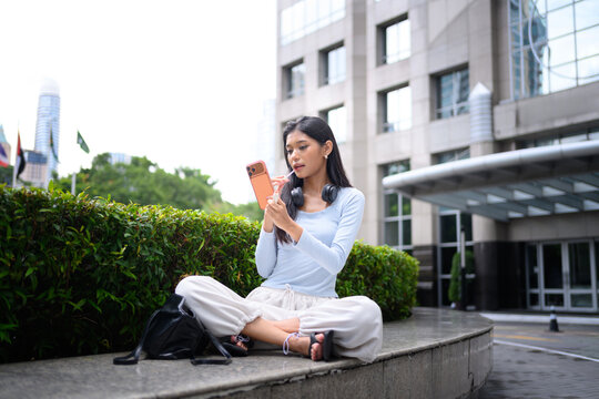 Young Asian Woman Sitting Outdoors in Urban Setting Browsing Smartphone with Headphones Around Neck