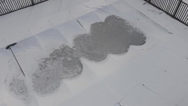 Overhead drone rotation reveals a snow covered fenced pool. A dark safety fence surrounds a white pool cover patterned with uneven patches of snow and thawed areas.