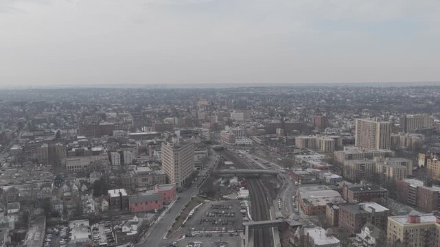 Wide drone pan across urban rail lines and apartment towers. Parallel tracks and road bridges cut through a dense mix of low rise buildings and taller apartment blocks.