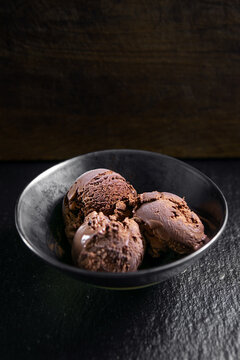 Rich chocolate ice cream scoops served in a black bowl on a dark textured surface, with negative space for advertising and copy
