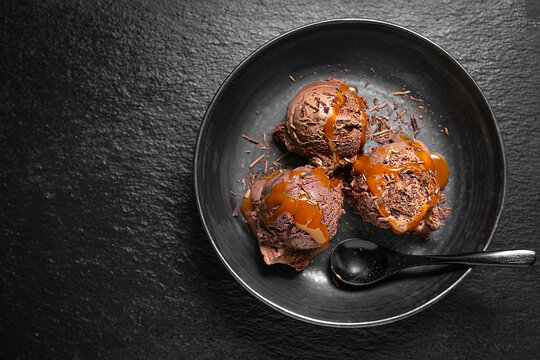 Rich chocolate ice cream scoops served with caramel sauce in a black bowl on a dark textured surface, with negative space for advertising and copy