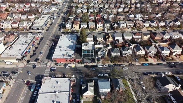 Drone tracks sideways over a crossroads and residential rooftops. The camera crosses the junction past flat roofed corner buildings and rows of detached houses. Filmed in Queens NYC.