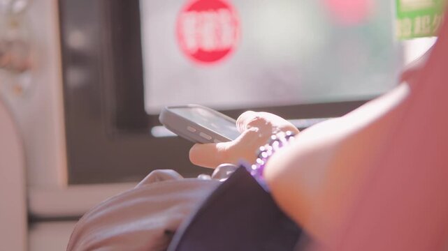 Hands lift and turn a dark smartphone beside a Taipei bus window in slow motion, with a pink shirt and beaded wrist bracelet visible as a passenger adjusts the device above a seated lap during travel.