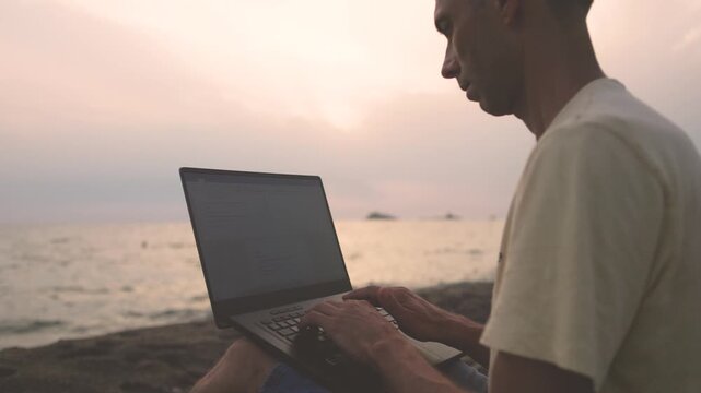 Side view of man working on laptop sitting on pebble beach at dusk
