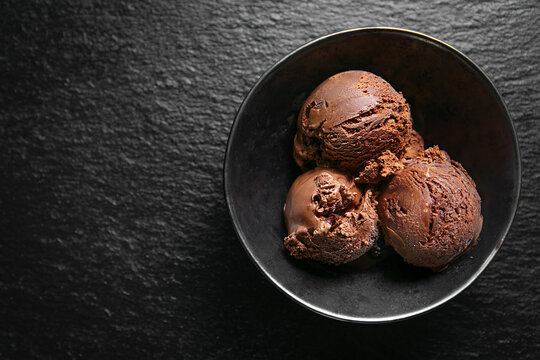 Rich chocolate ice cream scoops served in a black bowl on a dark textured surface, with negative space for advertising and copy