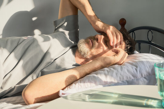 Man lying in bed suffering from headache, covering his eyes from sunlight