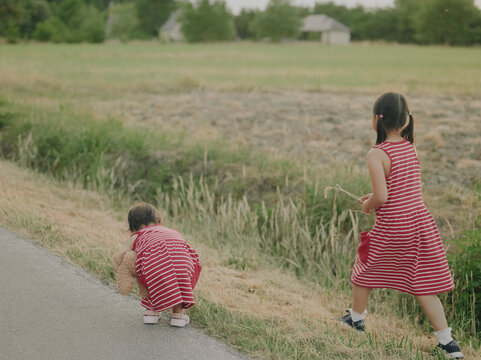 Two young sisters in red striped dresses walking away along a country road in the summer countryside of Pays de la Loire, France.