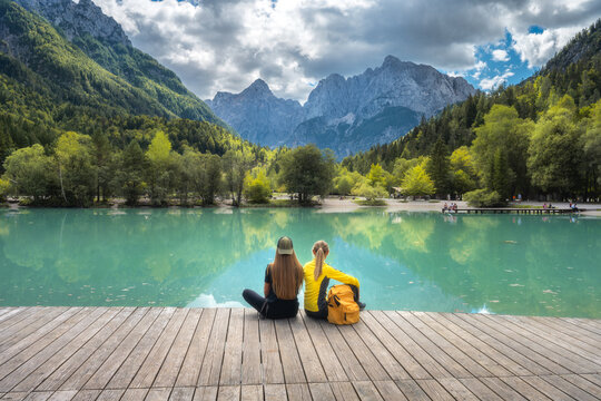 Two women sitting on wooden pier at Jasna Lake in Slovenia. Alpine landscape with turquoise water reflection, mountains, sky, clouds, green trees forest, yellow backpack and summer view. Autumn