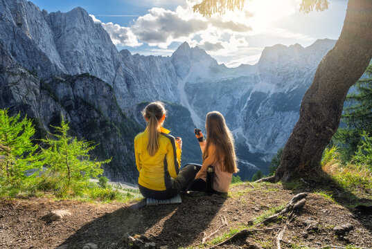 Two women hikers sitting under tree drinking tea in Slovenia. Alpine mountain landscape with rocky peaks, sky, clouds, sunbeams, sun flare, forest slope, thermos and autumn sunset view. Autumn alps