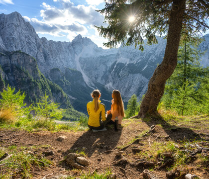 Two women hikers sitting under tree drinking tea in Slovenia. Alpine mountain landscape with rocky peaks, sky, clouds, sunbeams, sun flare, forest slope, thermos and autumn sunset view. Autumn alps