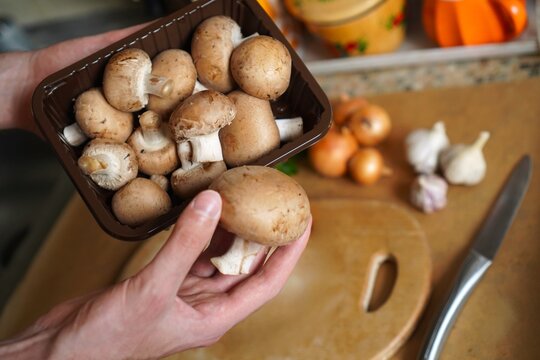 Hands holding pack of fresh brown champignon mushrooms over kitchen counter, knife and pumpkin in blur. Cooking vegetarian low-calorie dish with organic autumn ingredients, healthy food lifestyle.