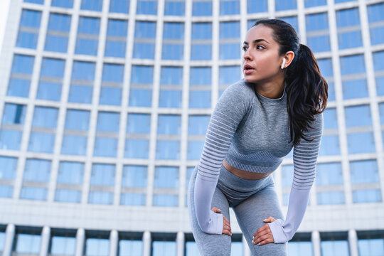 Close-up photo of 20s sportswoman resting after jogging in front of urban background, female athlete recovering outdoors, fitness lifestyle, running, wellness, healthy living active exercise concept