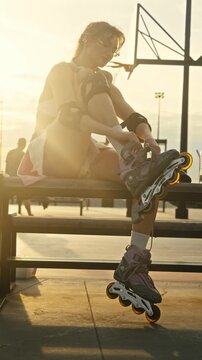 Tired woman unties inline skates on wooden bench after workout. Golden sunset light shines over urban extreme park while sportive female removes heavy sports gear.