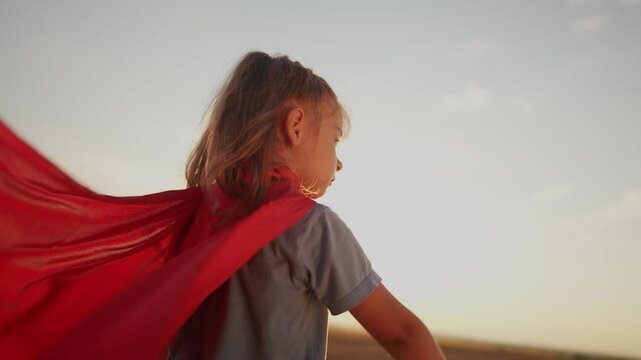 girl with the cloak. concept of happy childhood and family for a child. little girl in a superhero costume stretched hand forward, a red cape and a lifestyle blue T-shirt, sunset on the background
