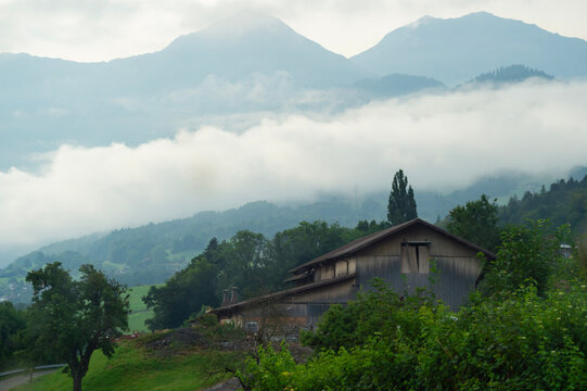 A wooden chalet stands on a green hillside in Lauterbrunnen, Switzerland. Low clouds and mist drift around forested mountain peaks in the background.