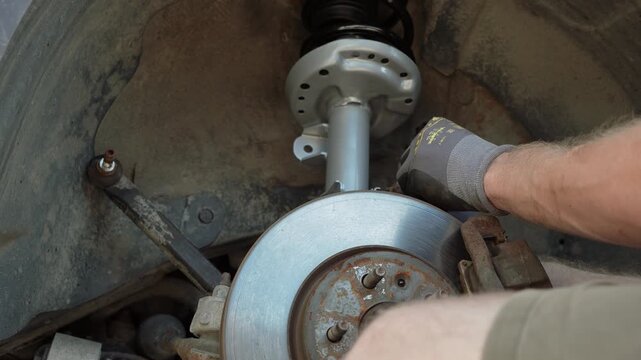 Mechanic hands in gloves installing a new silver front suspension strut shock absorber with coil spring into a car wheel well during a repair process in a workshop, close up of car maintenance. 